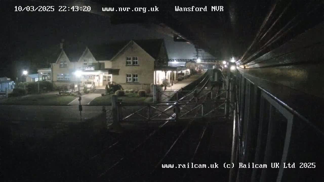 A nighttime view from a train shows a small, illuminated building next to a train station, with train tracks extending into the darkness.