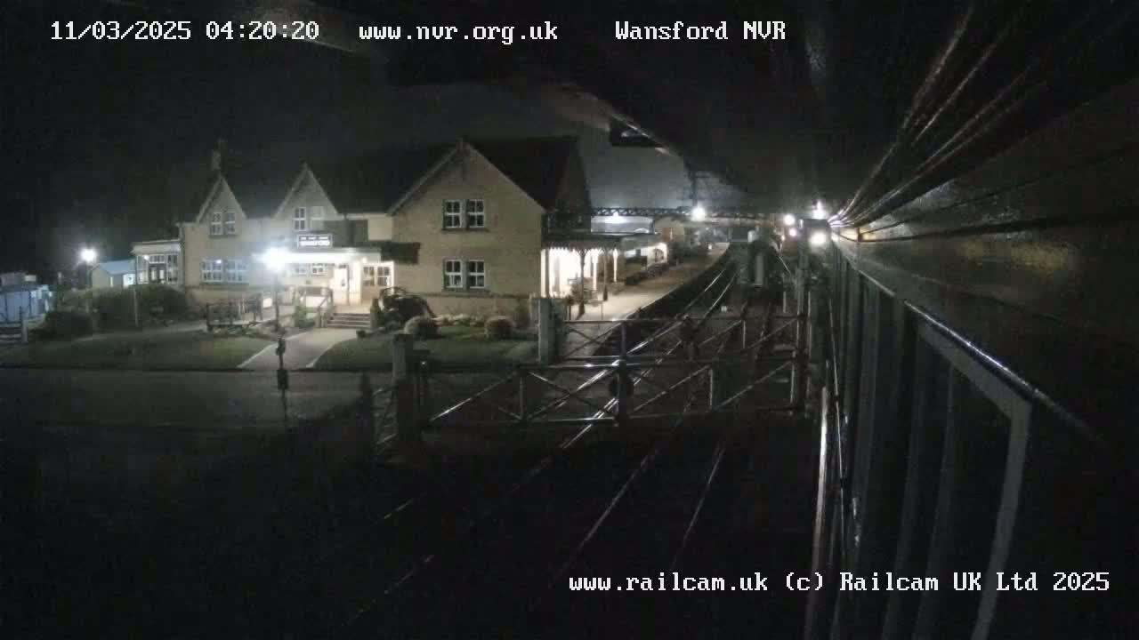 A nighttime view from a train shows a two-story building with lights on, next to a train station platform and tracks.