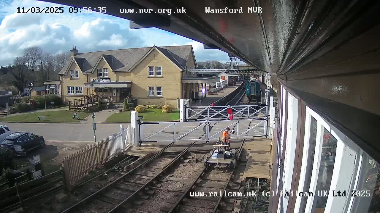 A partly cloudy day at a train station shows a railway crossing with a person working on a small cart near a railway and a building in the background.