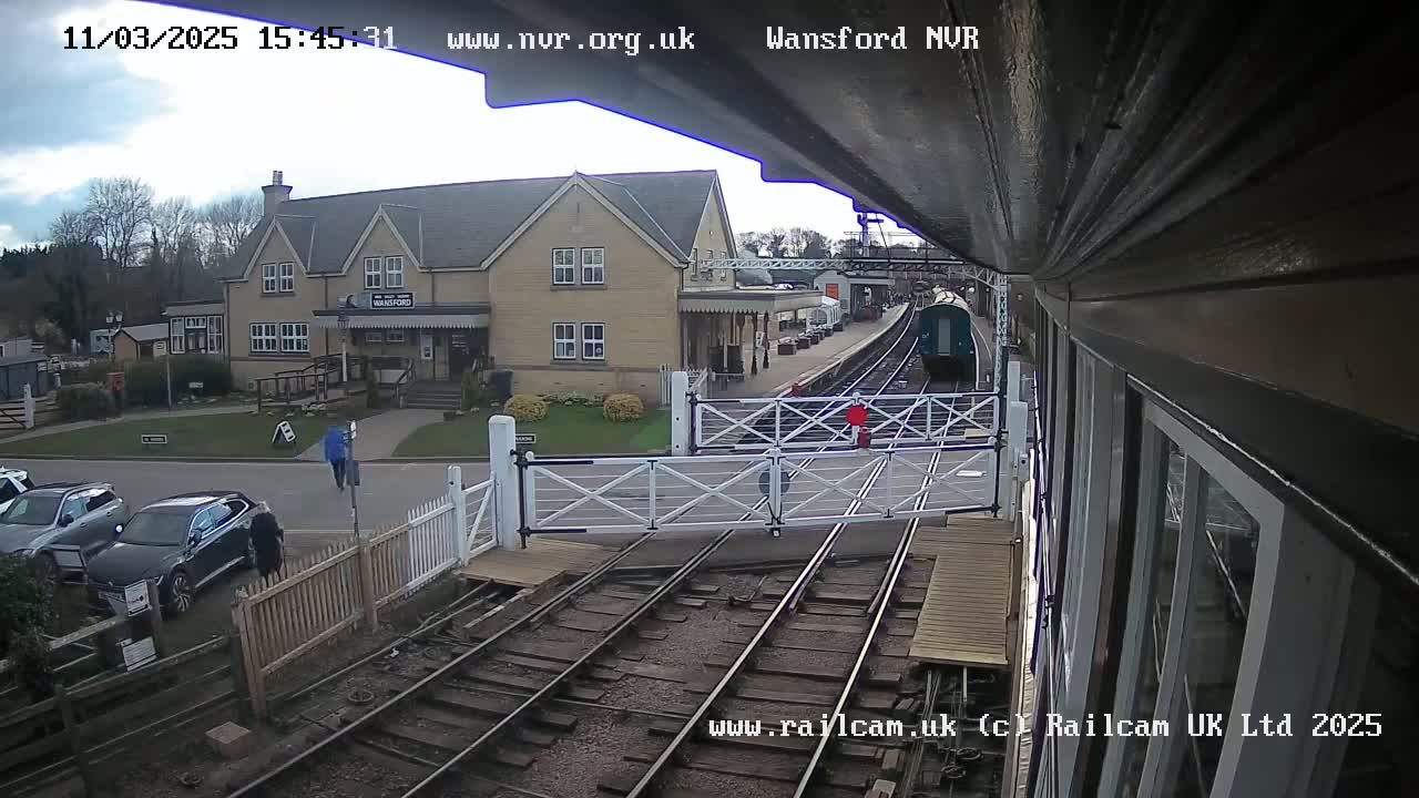 A train approaches a railway crossing near a station building on an overcast day.