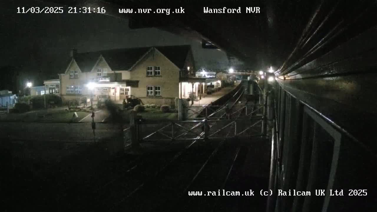 A nighttime view of a train station with a two-story building, illuminated by artificial lights, under dark skies.