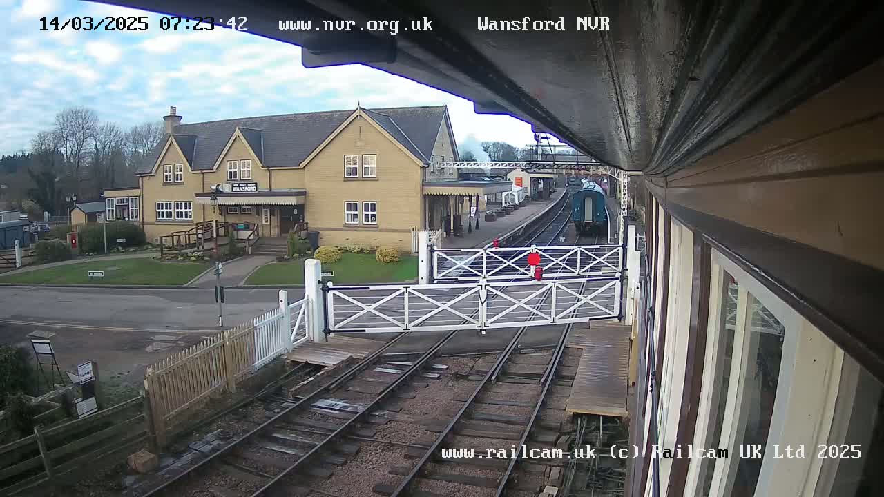 A train approaches a station on a partly cloudy day, passing through a level crossing gate in front of a yellow building.