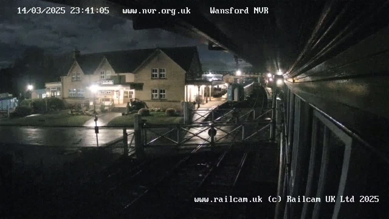 A nighttime view of a train station with a building and a train partially visible under a cloudy sky, with wet pavement.