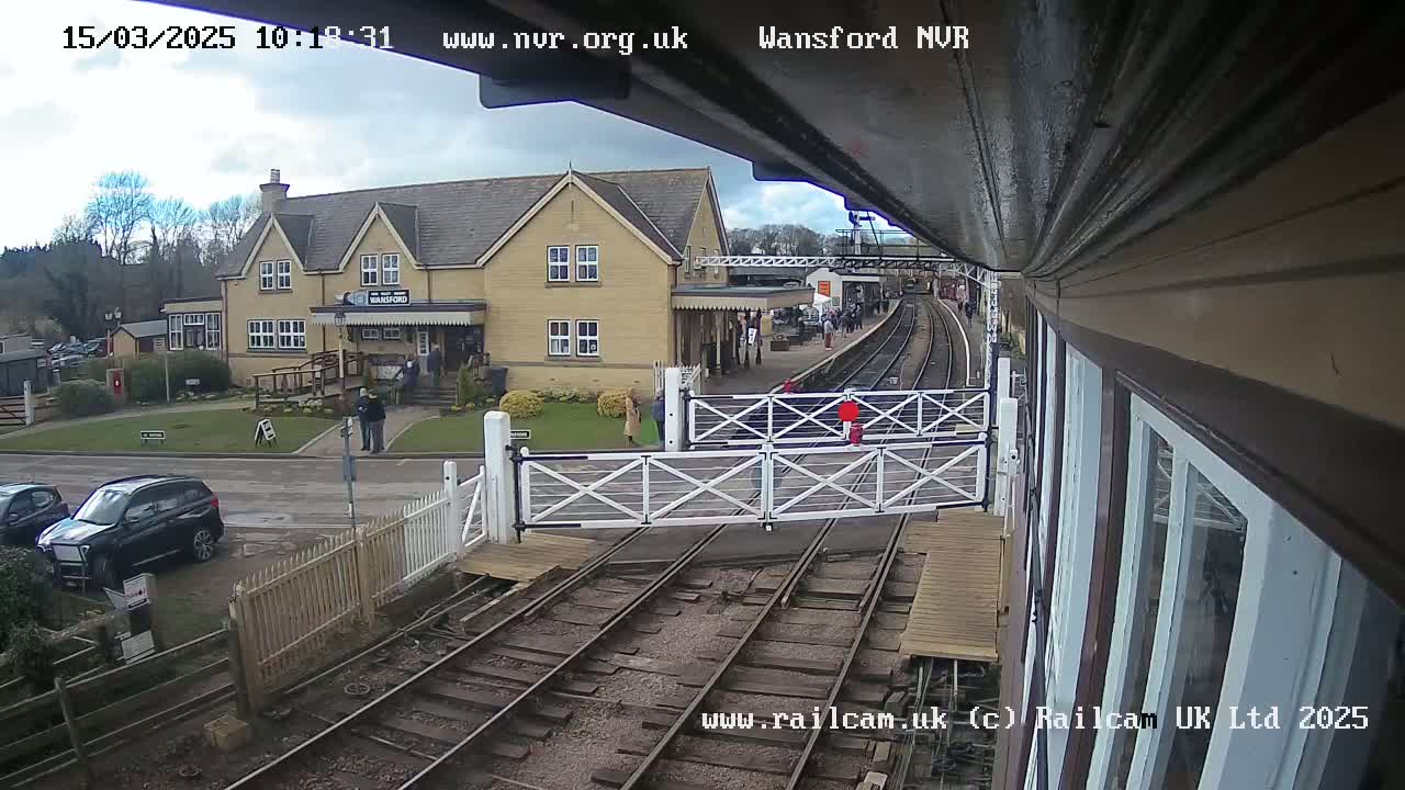 A partly cloudy day at a train station with a building and a few people visible, viewed from a slightly elevated position near a level crossing.