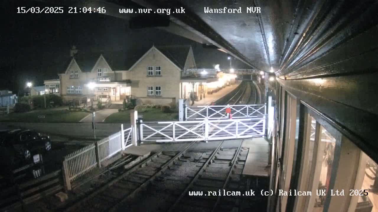 A nighttime view from a low angle shows a train station with a two-story building and crossing gates closed over railroad tracks.