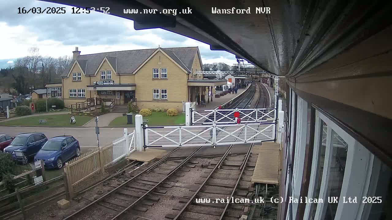 A railway station with a two-story building and a partly cloudy sky is viewed from a train car.