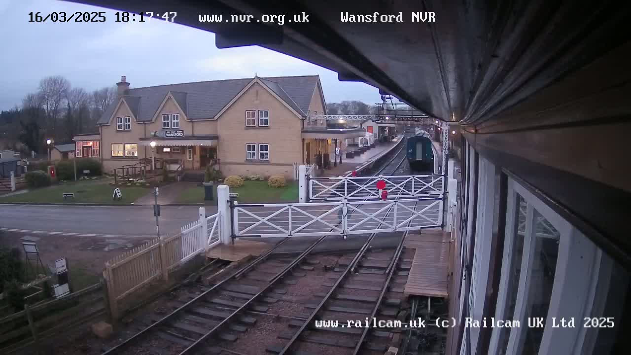 A train approaches a station on an overcast day, passing through a level crossing with white gates.