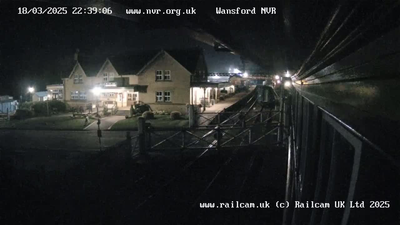 A nighttime view from a train shows a station building and platform illuminated, with a dark, overcast sky.