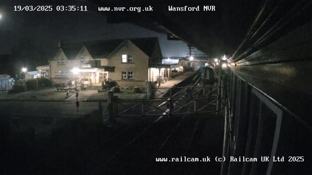 A nighttime view from a train shows a small, lit station building and train tracks.