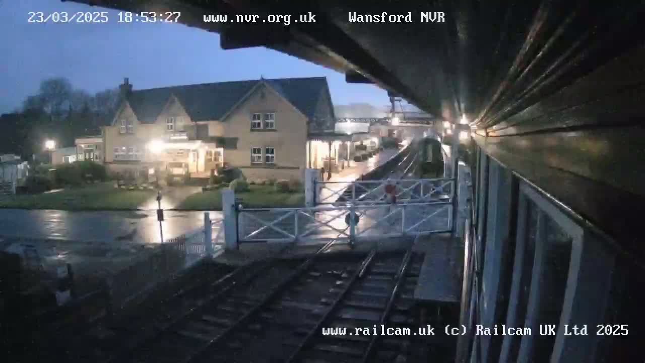 A wet nighttime view from a train shows a two-story building with lights on, a railway crossing, and train tracks extending into the distance.