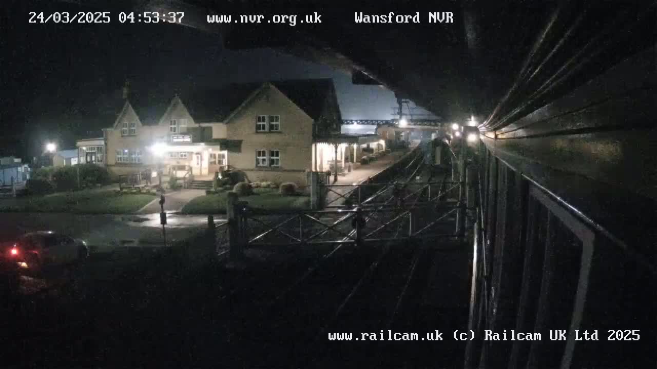 A nighttime view of a train approaching a station building next to a road with a car, under dark, clear skies.