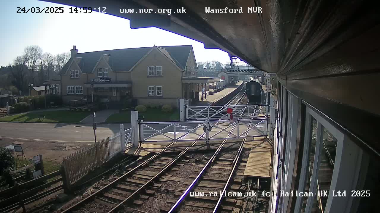 A train approaches a railway station on a sunny day, passing through a level crossing in front of a stone building.