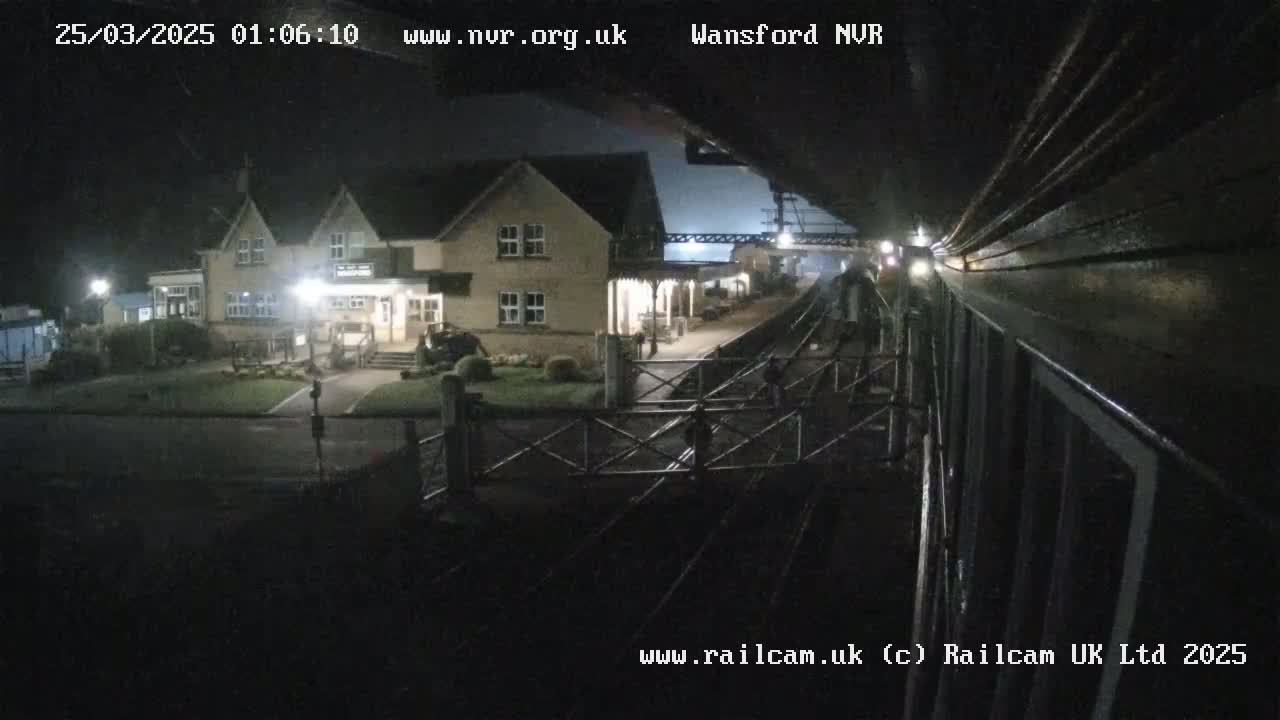 A nighttime view from a train shows a station building and platform, with a level crossing gate and tracks in the foreground under a dark sky.