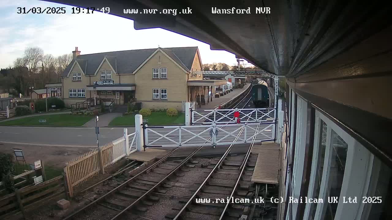 A train sits at a station on a partly cloudy evening, with level crossing gates down across a road in front of a light-colored building.