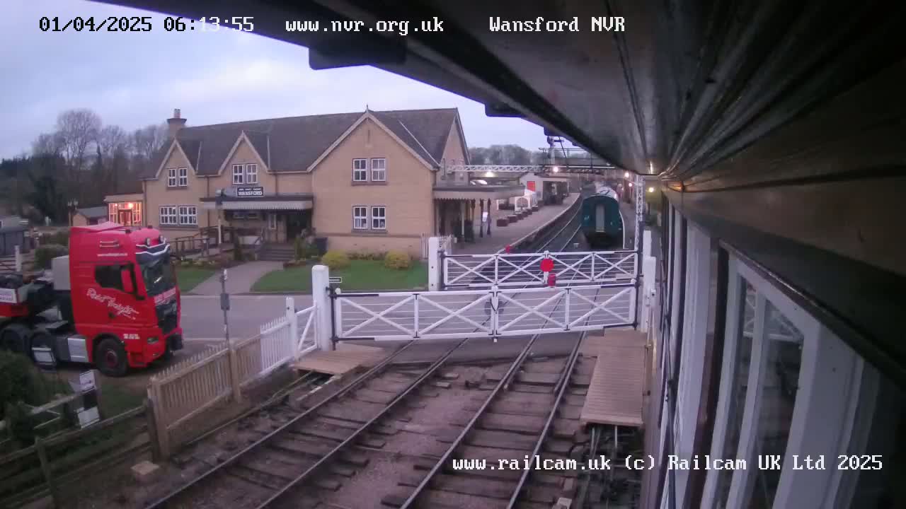 A red semi-truck is parked near a railway crossing gate, with a train partially visible in the background on an overcast day.