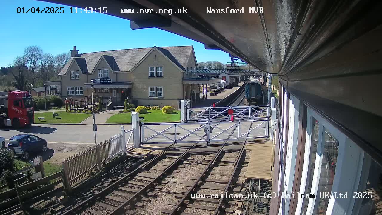A train is approaching a railway station on a sunny day, with a red truck and cars visible nearby.