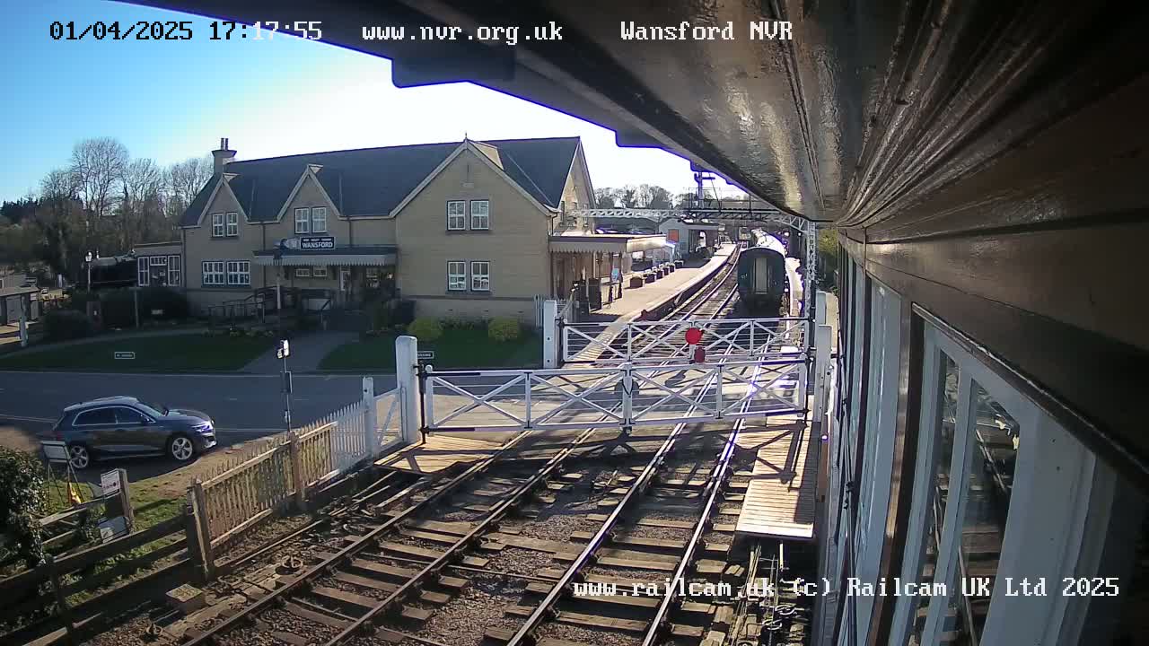 A train is arriving at a railway station on a sunny day, with a car parked nearby and a level crossing gate down.