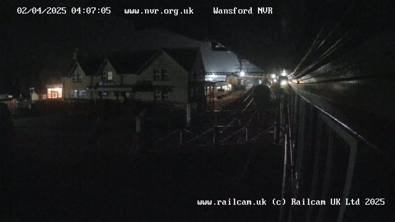 A nighttime view of a train approaching a station, passing a building with lights on, under dark, cloudy skies.