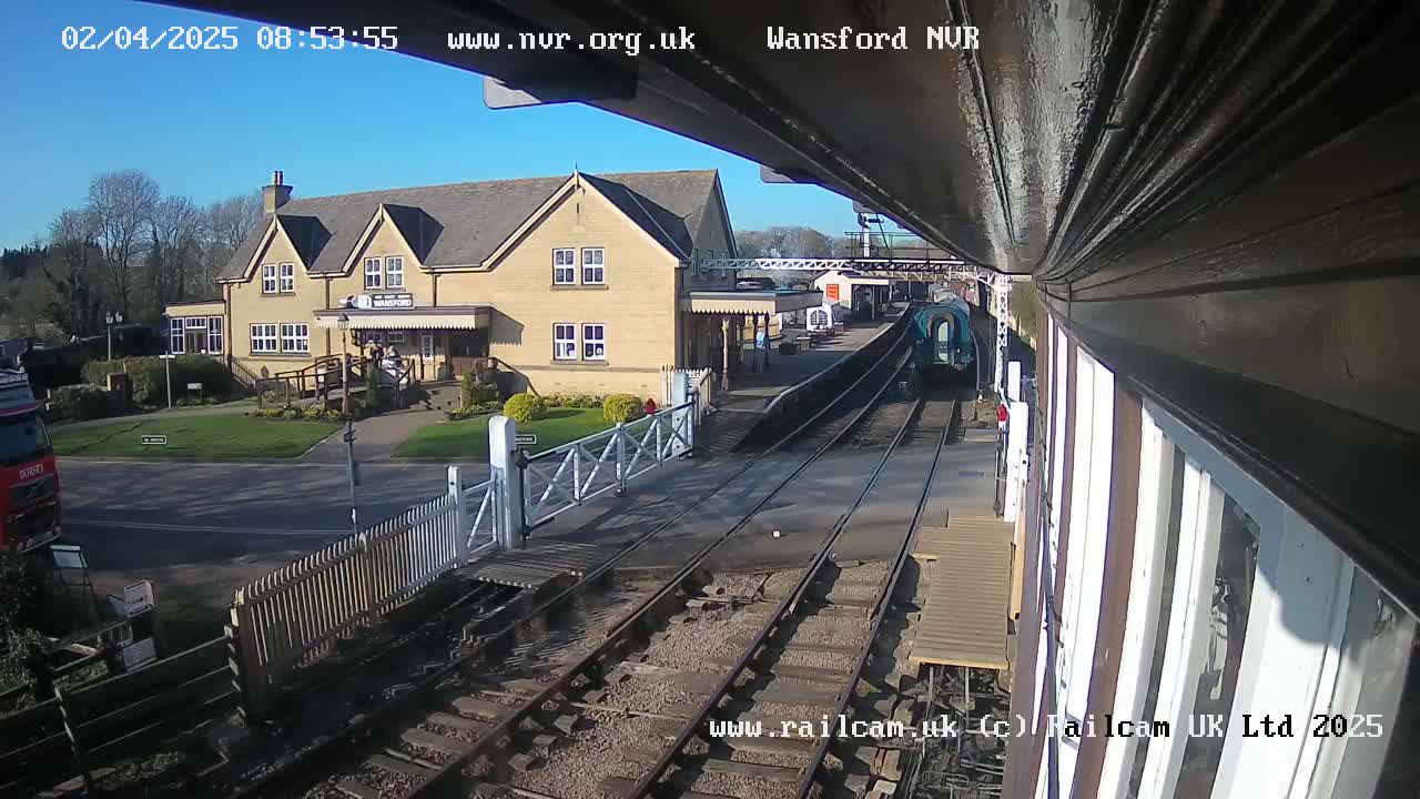 A train departs from a station on a sunny day, passing a yellow building and a level crossing.