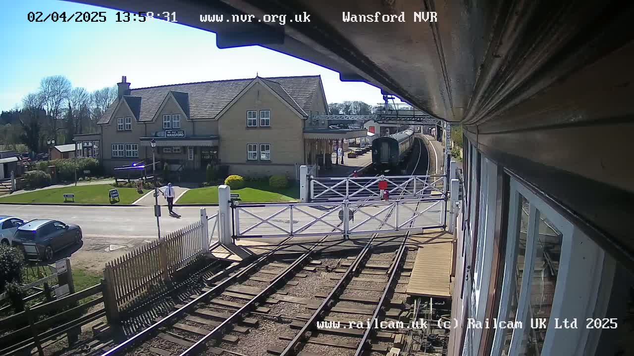 A train approaches a station on a sunny day, passing through a level crossing with cars parked nearby and a person walking in the foreground.