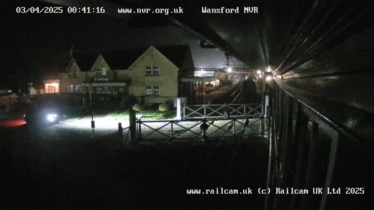 A nighttime view of a train approaching a station with a building and level crossing visible, under dark skies.