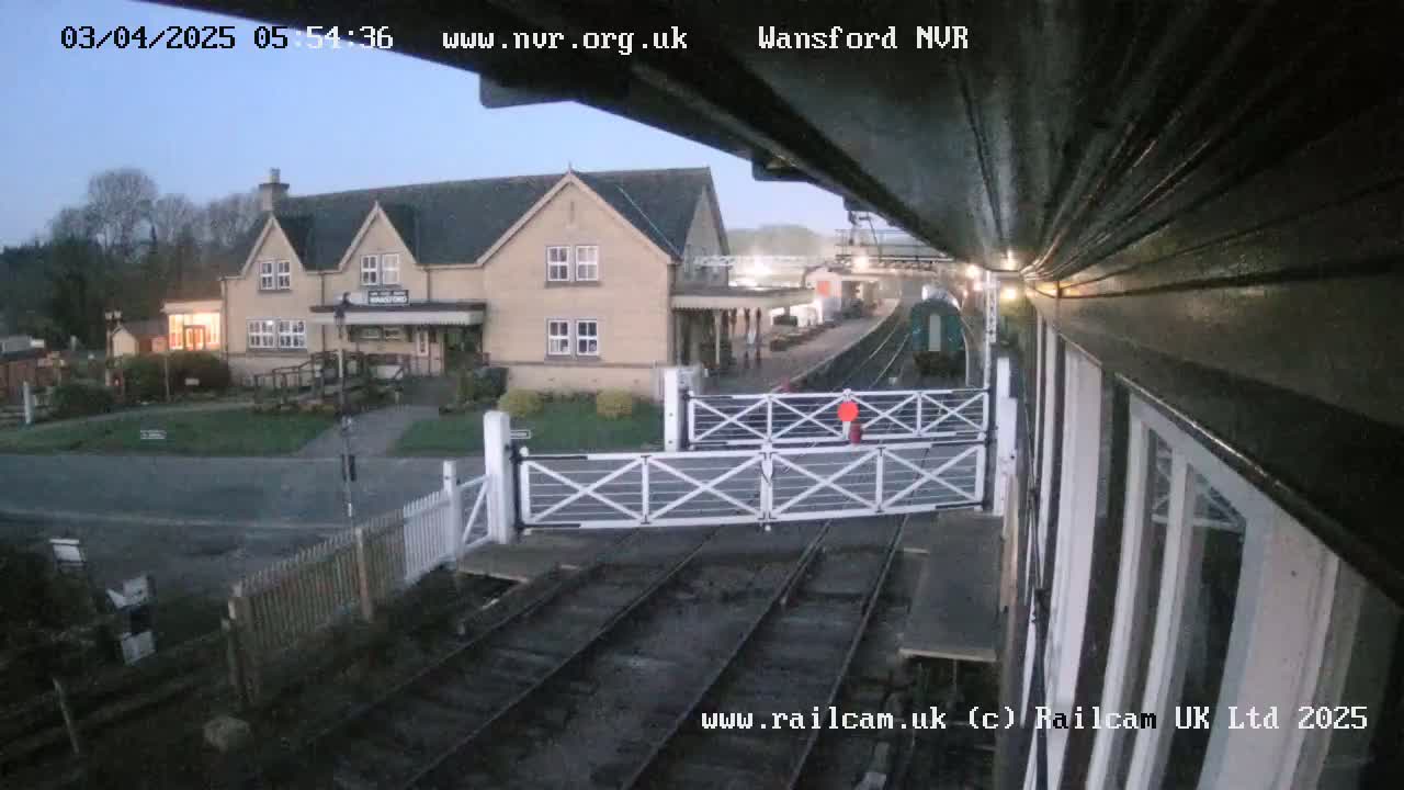 A train passes through a railway crossing near a building in the early morning under a twilight sky.