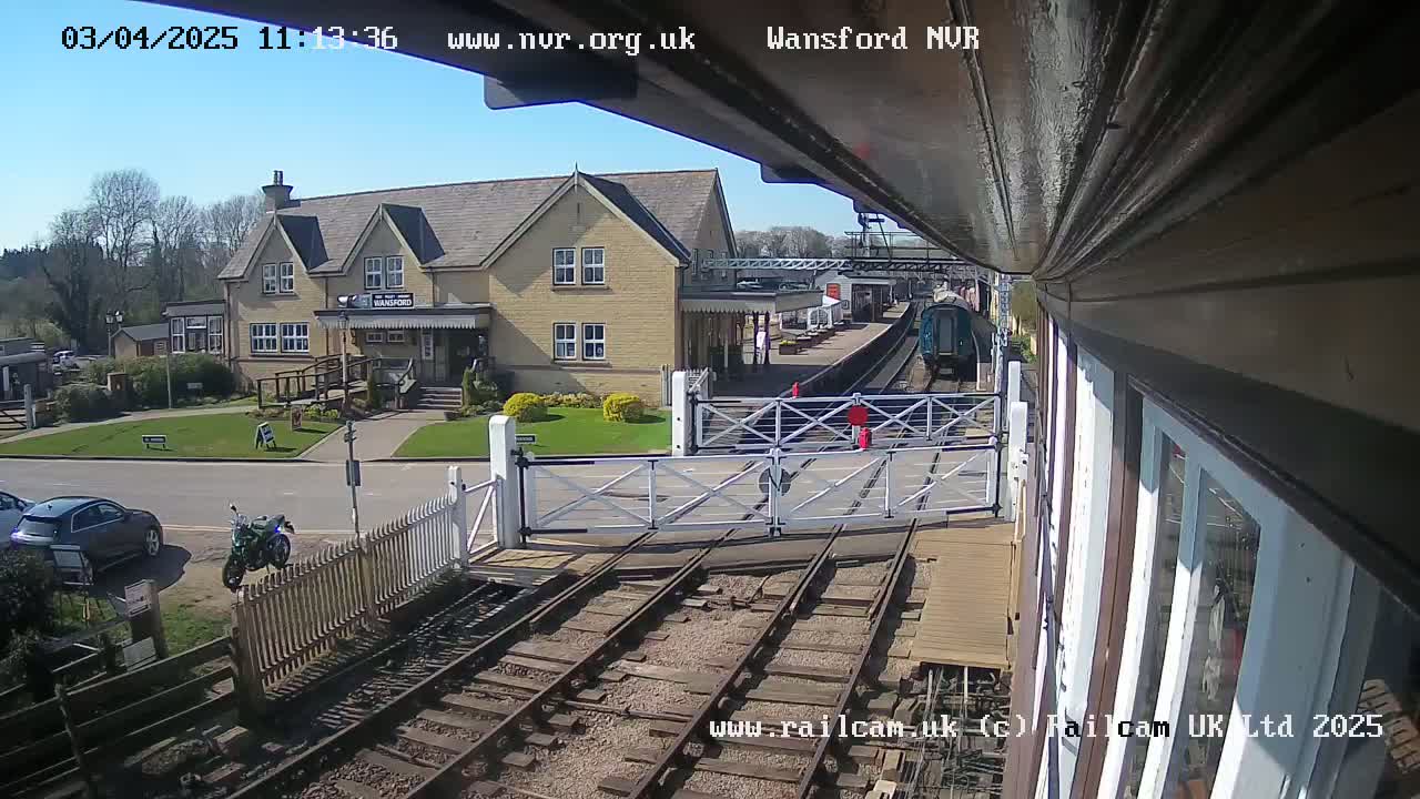 A train is at a station on a sunny day, with a two-story building visible nearby and cars and a motorcycle parked in the area.