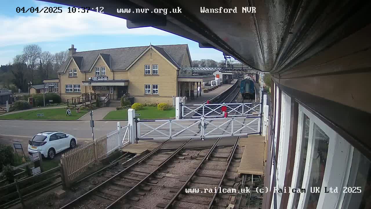 A train is arriving at a railway station on a partly sunny day, with a white car parked nearby and level crossing gates in the down position.