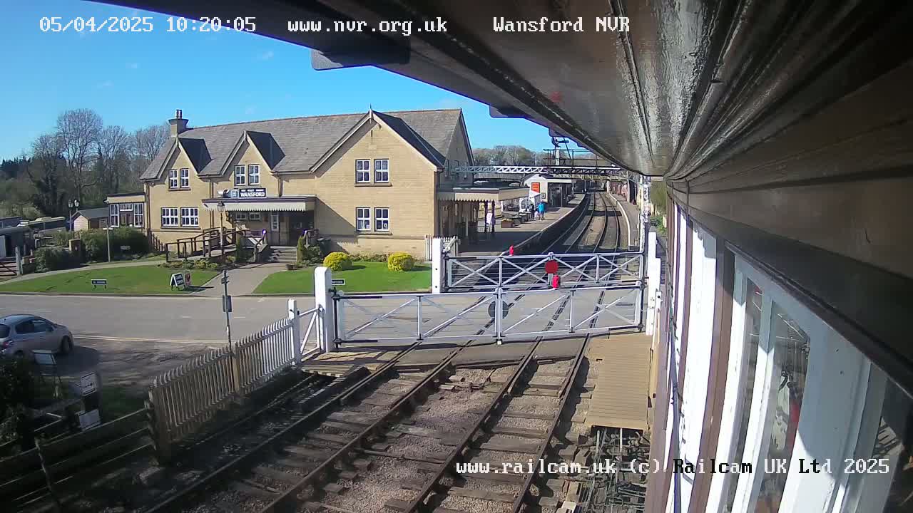 A sunny view of a train station with a two-story building, train tracks, and a level crossing gate.