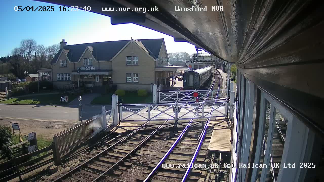 A railway crossing with a train approaching a station building on a sunny day.