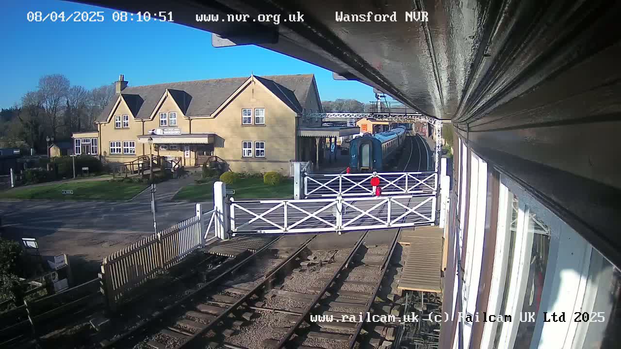 A blue train is passing through a railway crossing near a two-story building on a sunny day.