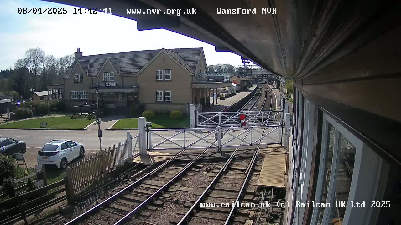 A view from a train shows a sunny day at a train station with a building, cars parked nearby, and a level crossing gate.