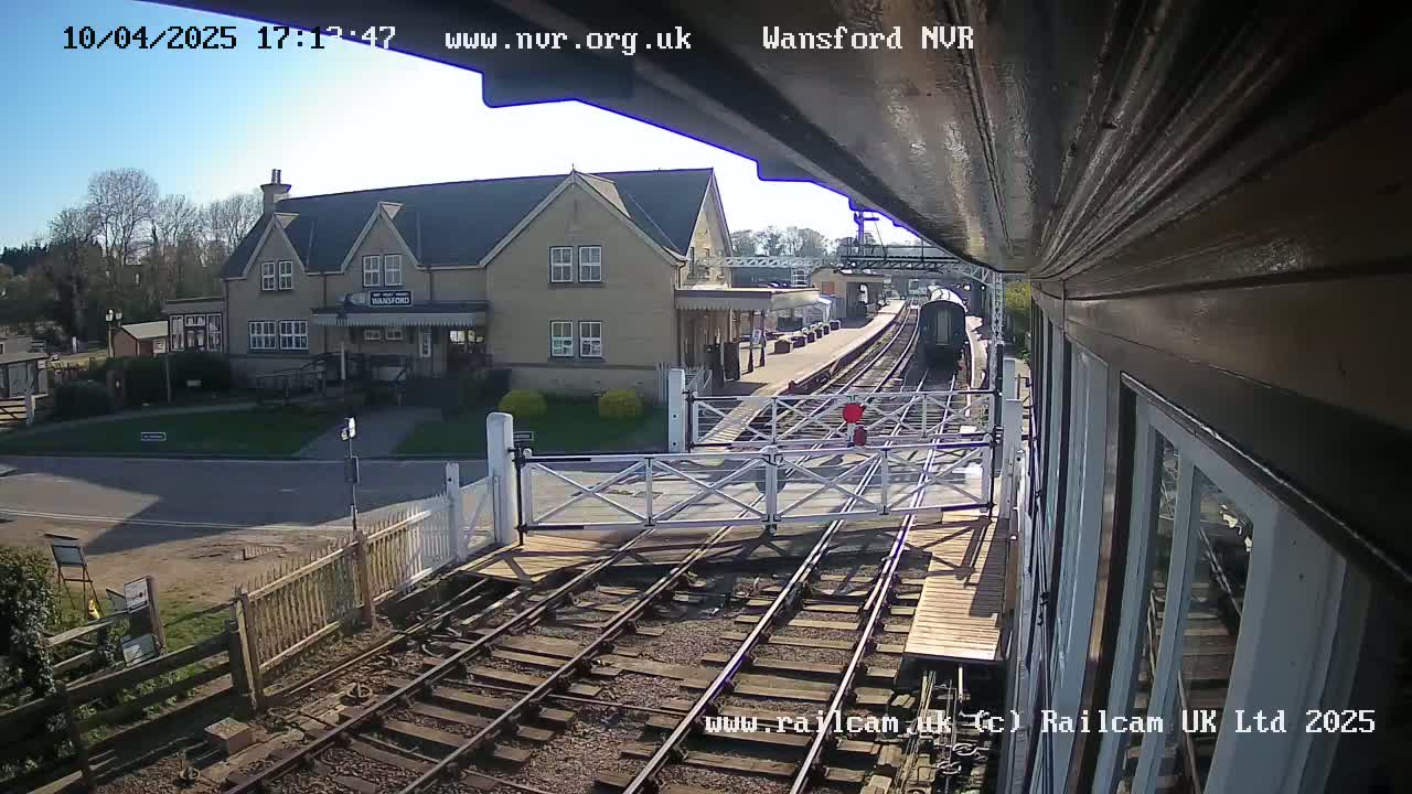 A train approaches a station on a sunny day, passing through a level crossing in front of a two-story building.