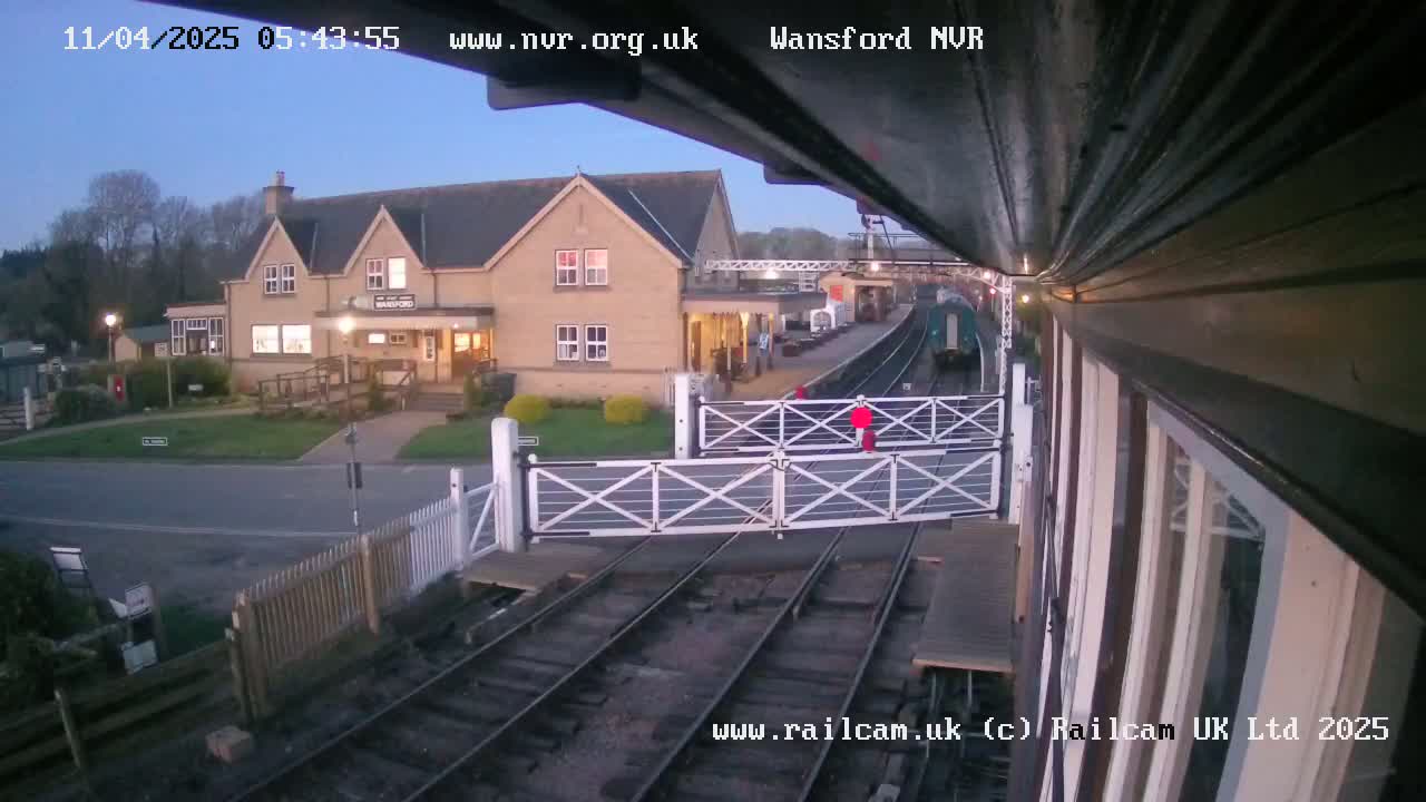 A train is approaching a station building under a twilight sky, with level crossing gates down.