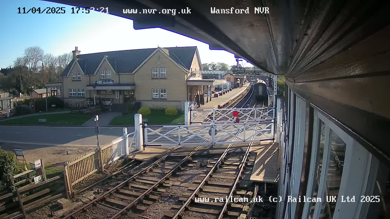 A train approaches a railway station on a sunny day, with level crossing gates down.