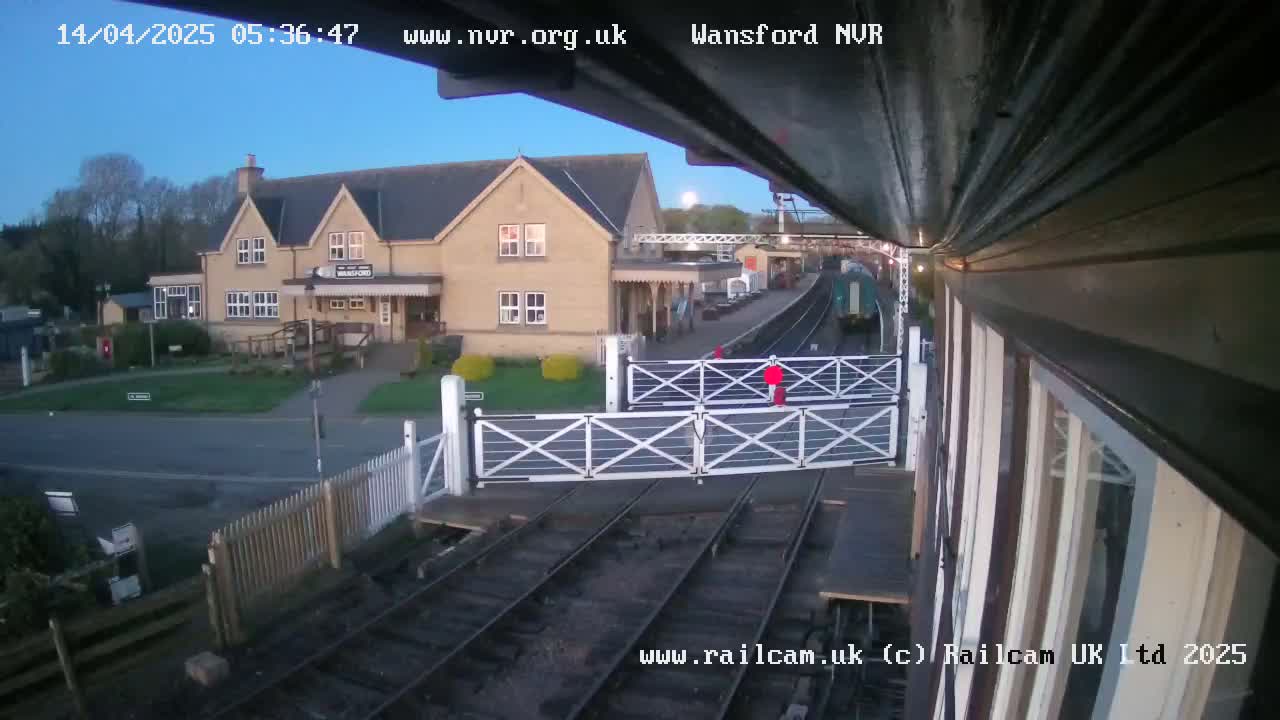 A train passes a railway station building on a clear morning, with level crossing gates down.