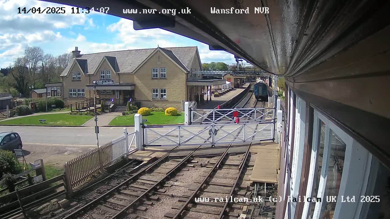 A train departs a station on a sunny day, passing through a level crossing near a light-colored building.