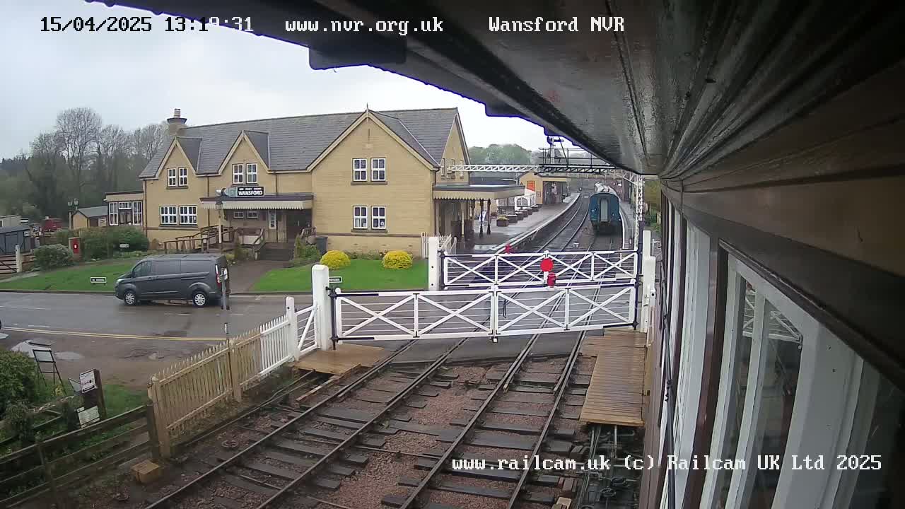 A train approaches a station on an overcast day, passing through a level crossing gate near a building and a van.