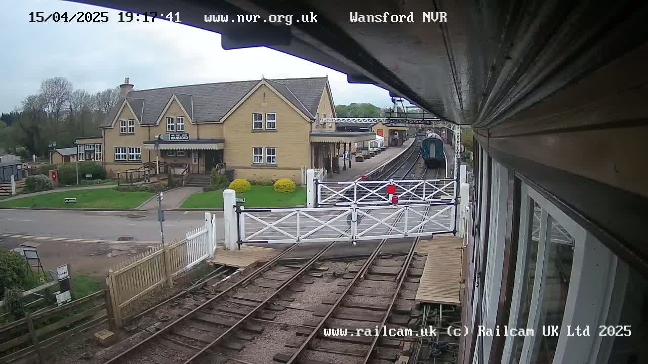 A train approaches a station on an overcast day, passing through a level crossing gate in front of a light-colored building.