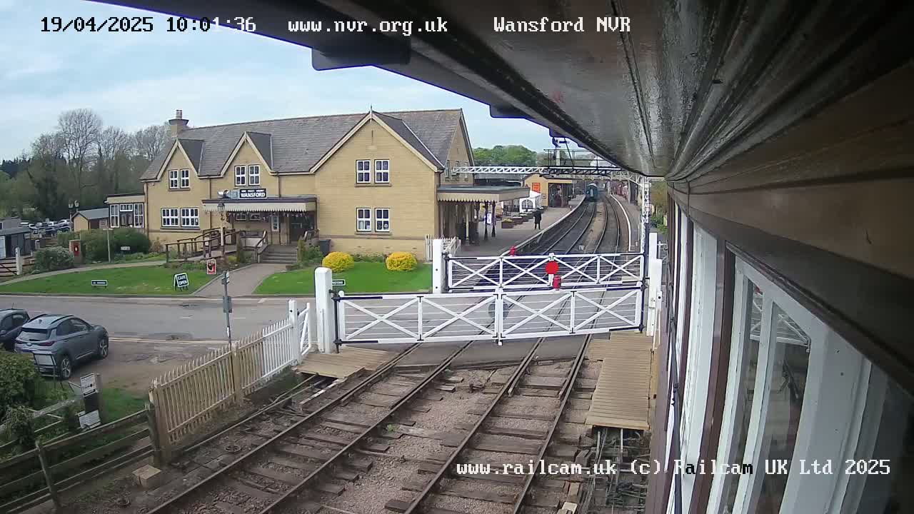 A view from a train station's signal box shows a partly cloudy day, a train on the tracks, a building with a station sign, and a level crossing with its gates down.