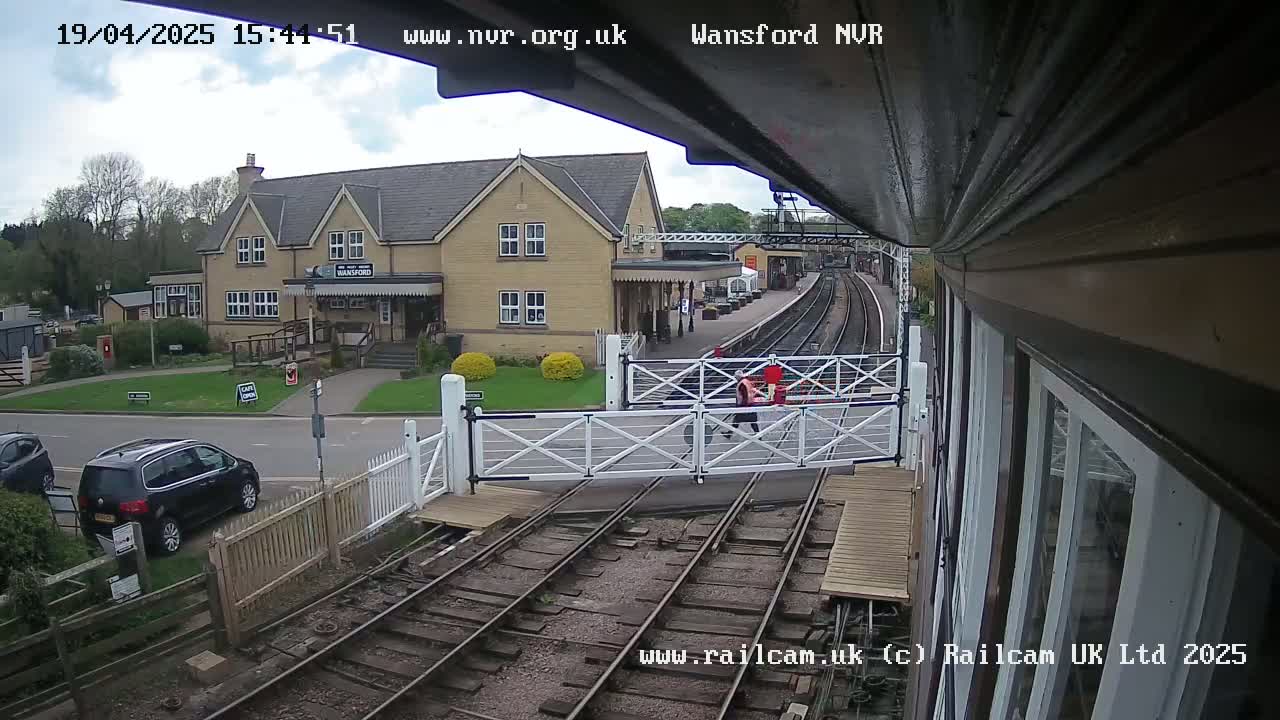 A partly cloudy day at a train station shows a building, railway tracks, a level crossing gate, and several parked cars.