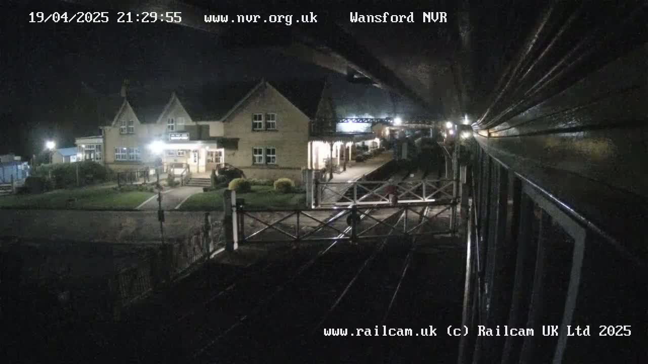 A nighttime view from a train shows a two-story building and a train station platform with a level crossing gate.