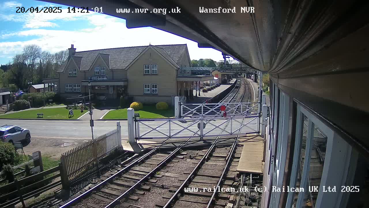 A train station with a two-story building, a road, and railway tracks is visible under a mostly sunny sky.