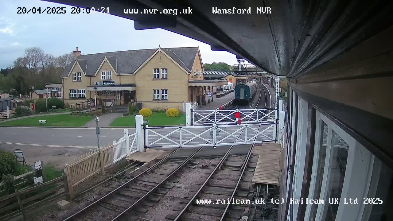 A train approaches a station building on a partly cloudy day, passing through a level crossing.