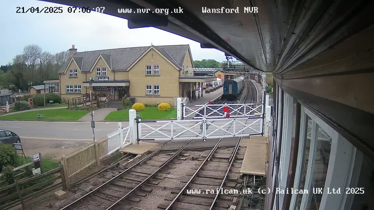 A train is arriving at a station on a partly cloudy day, passing through a level crossing in front of a light-colored building.