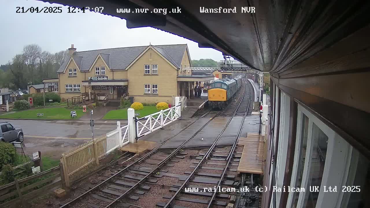 A green diesel train pulls into a wet train station next to a two-story building.