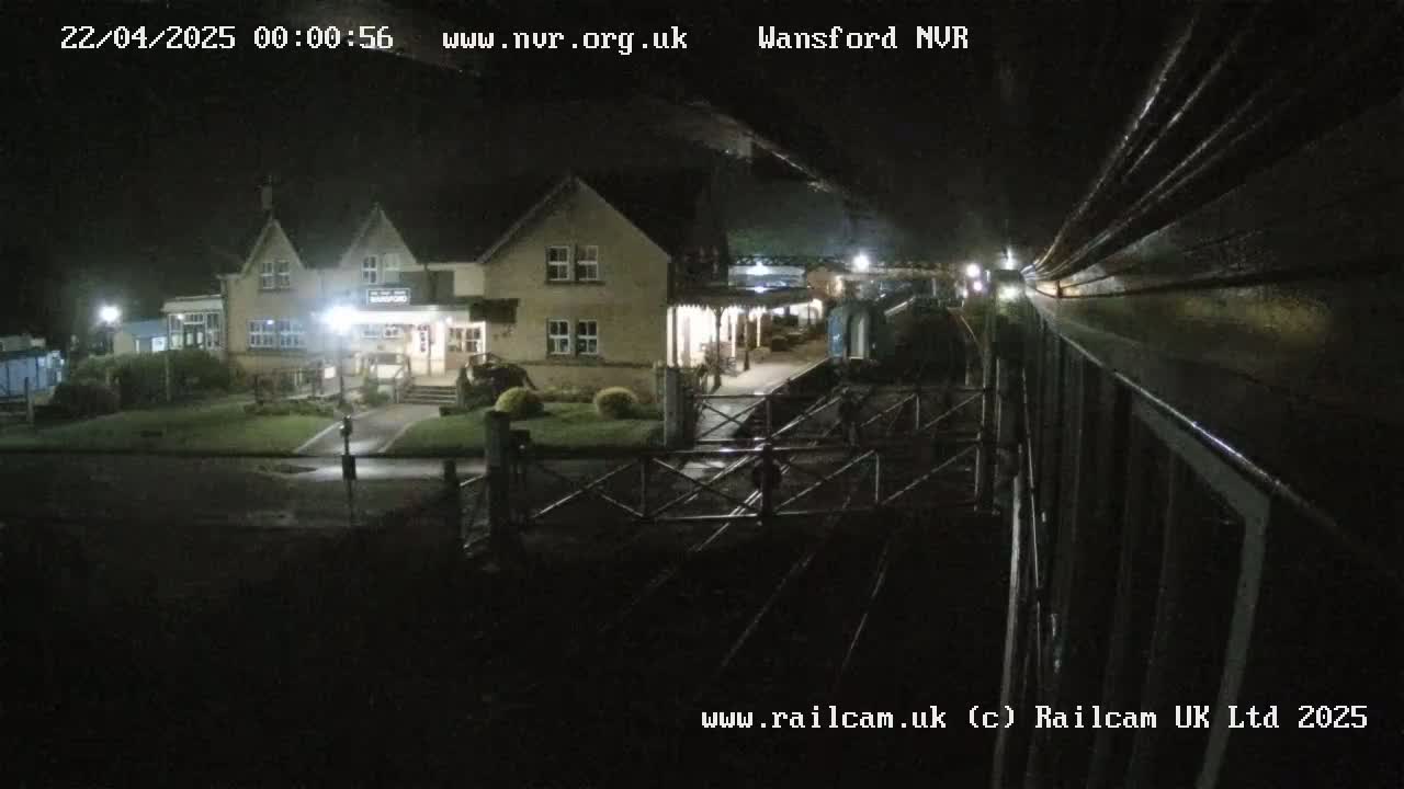 A nighttime view from a train shows a station building with lights on, and a section of railway tracks in the foreground.
