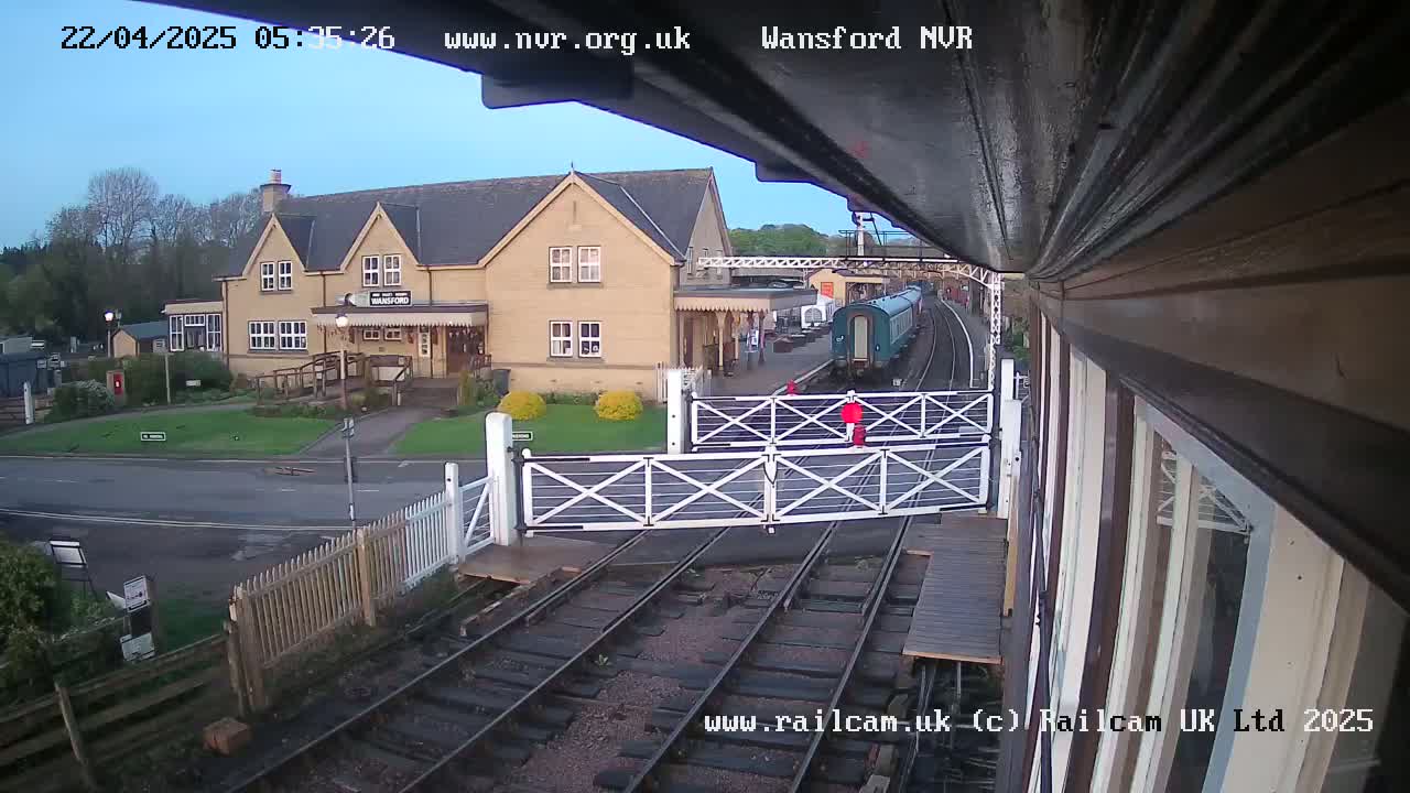 A train is departing a station on a partly cloudy morning, passing through a level crossing in front of a stone building.