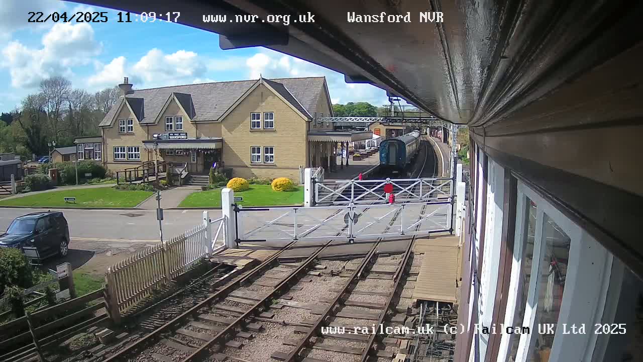 A train is arriving at a station on a partly sunny day, with a building and a vehicle visible in the foreground.
