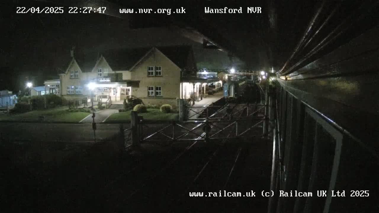 A nighttime view from a train shows a two-story building with lights on, next to a railway station under a dark sky.